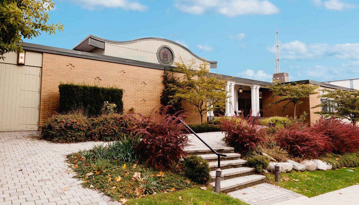 A single-story brick building with a curved roofline, stone entrance steps, and landscaped bushes under a blue sky.