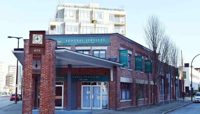 A corner brick building on a street with a clock tower feature, signage for funeral services, and trees along the side.