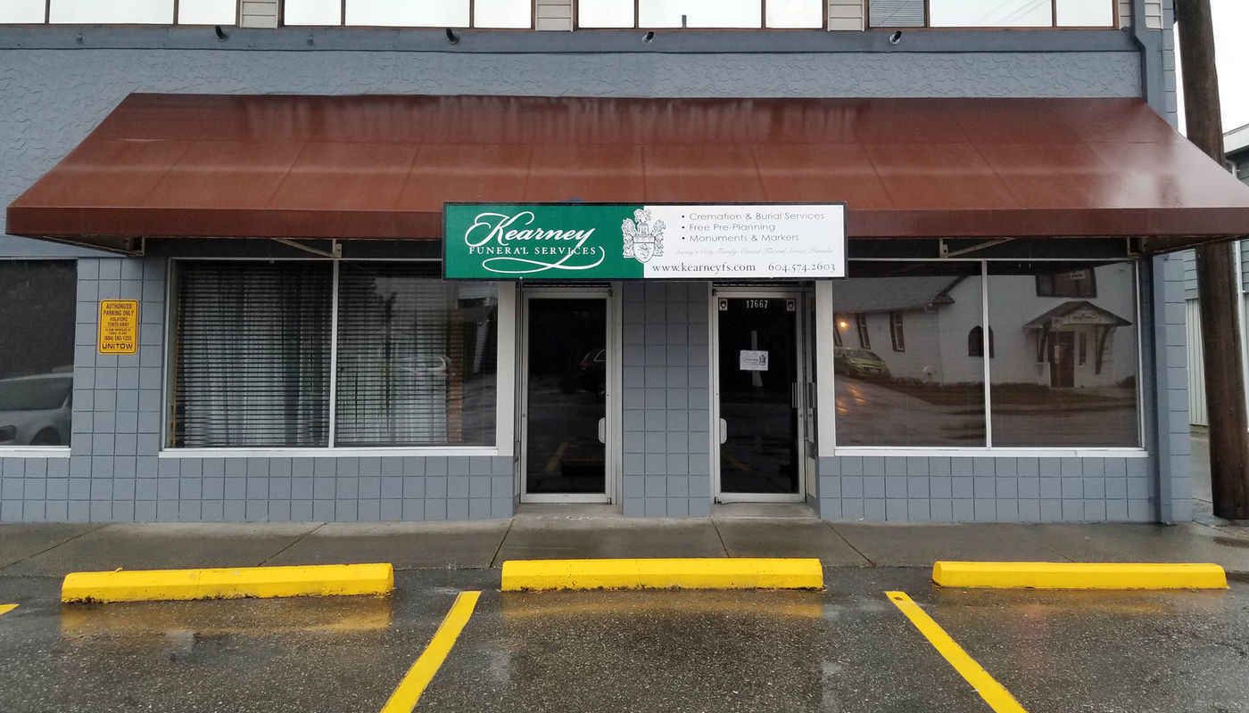 The front of a grey commercial building with a brown awning, a green sign, and three yellow parking bumpers.