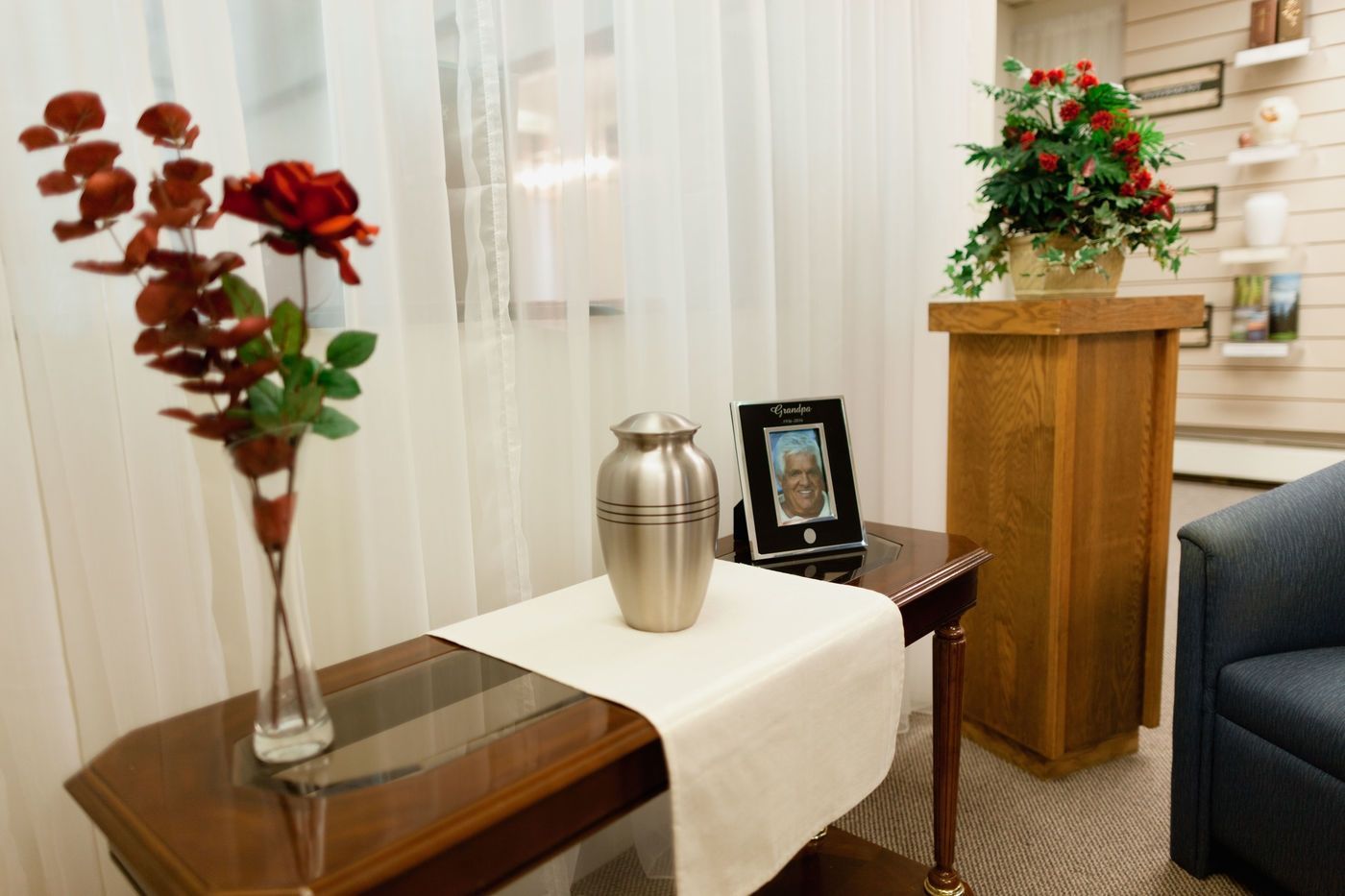 A memorial display with a metal cremation urn and a framed photo on a table, next to a wooden pedestal with flowers.