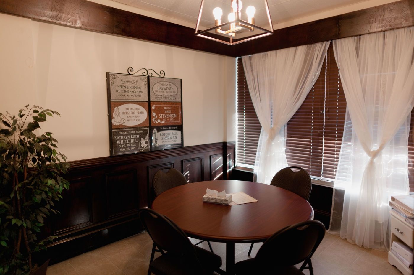 A round wooden table with four chairs sits in a room with dark wood paneling, white walls, and a light fixture above.