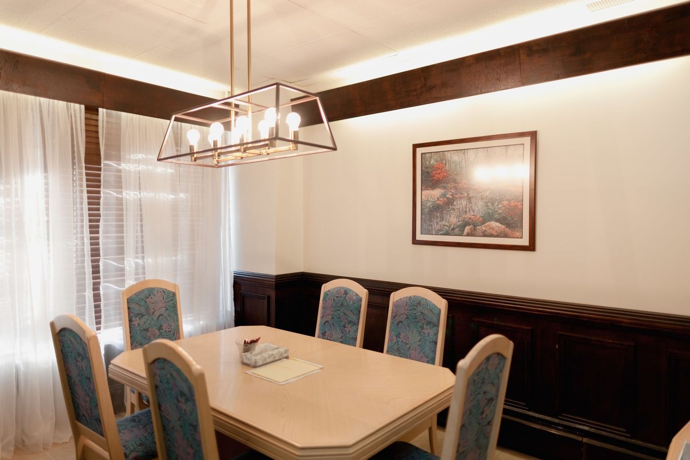 A dining room with a rectangular table, six blue-patterned chairs, a metal pendant light, and framed art on white walls.
