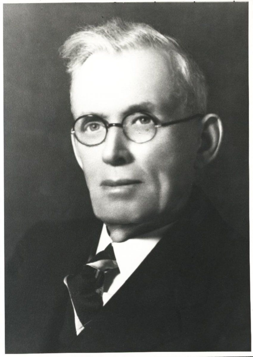 Black and white portrait of a man wearing glasses, a collared shirt, tie, and suit jacket against a dark background.