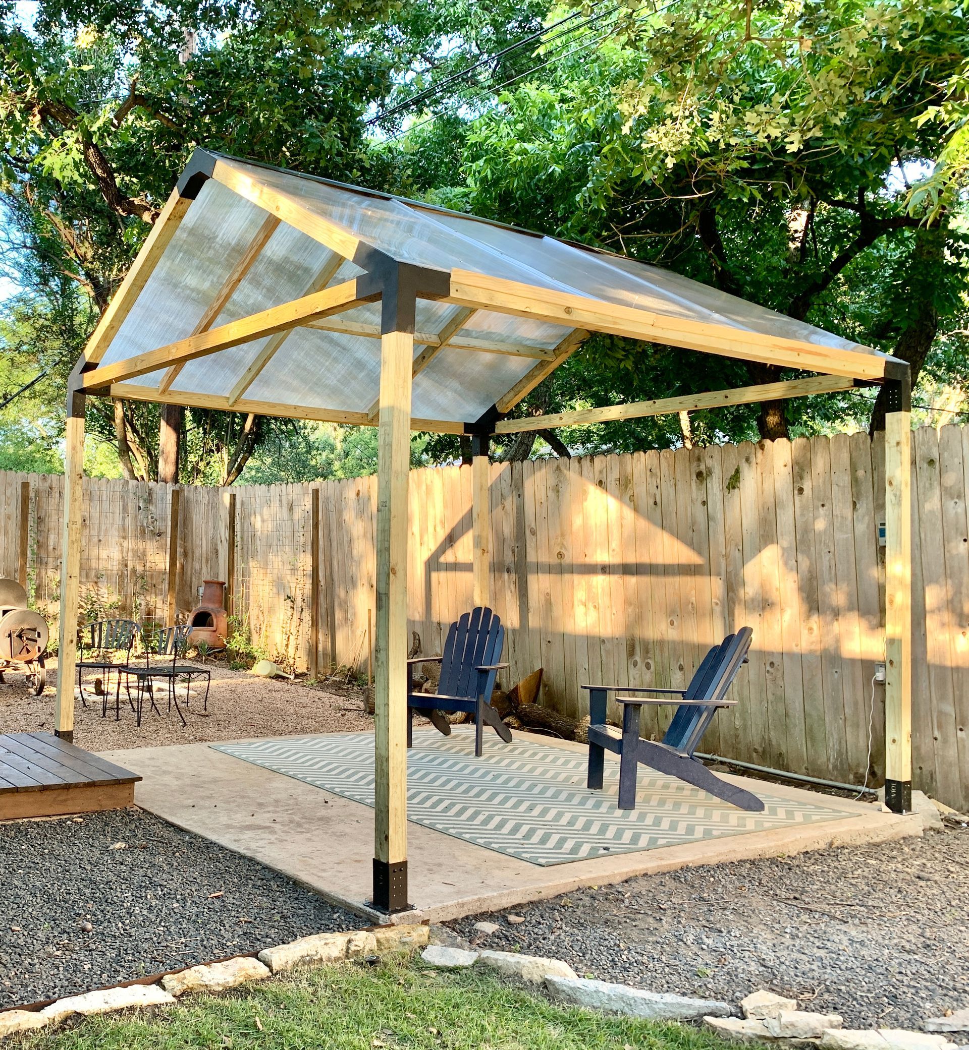 Backyard patio with a small metal-roof canopy, two chairs, and a wooden fence surrounded by gravel and trees.