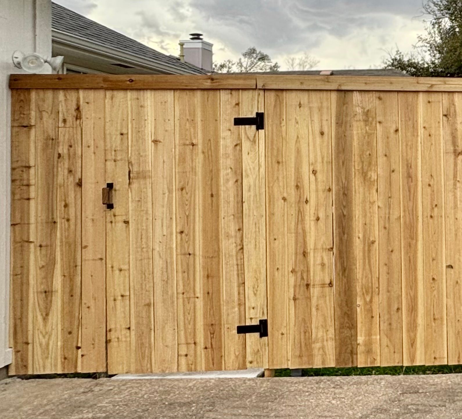 Wooden driveway gate with black hinges and latch beside a house, under an overcast sky