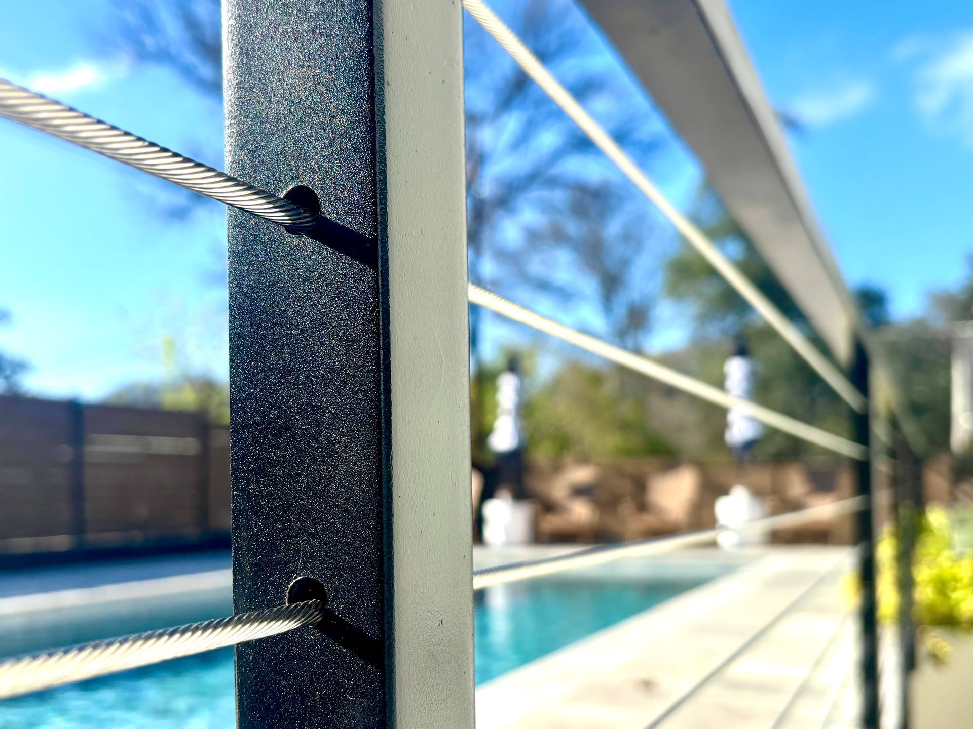 Close-up of cable railing beside a sunlit pool with a blurred backyard in the background
