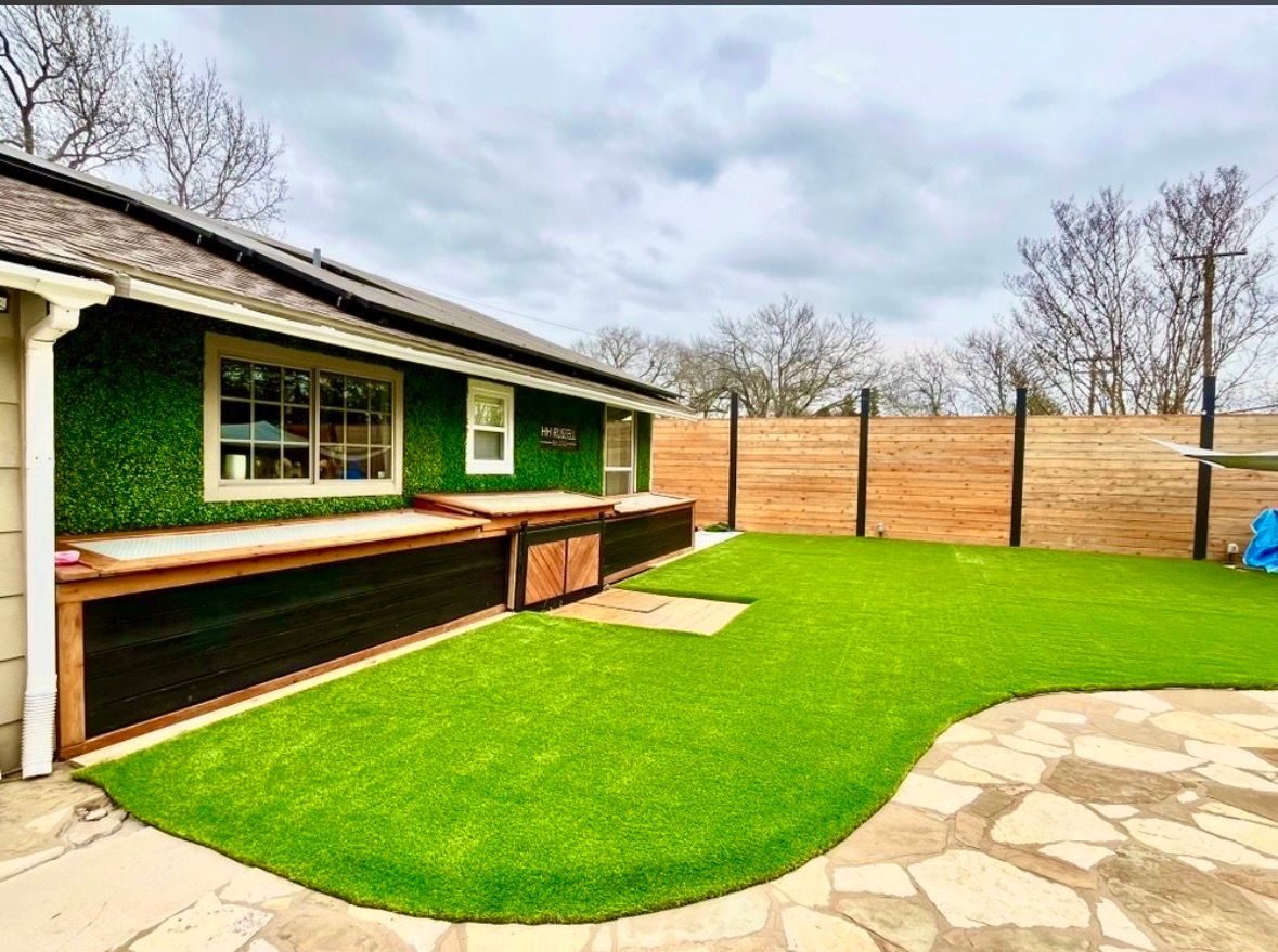Backyard patio with green lawn, stone path, wooden deck, and tan fence beside a house.