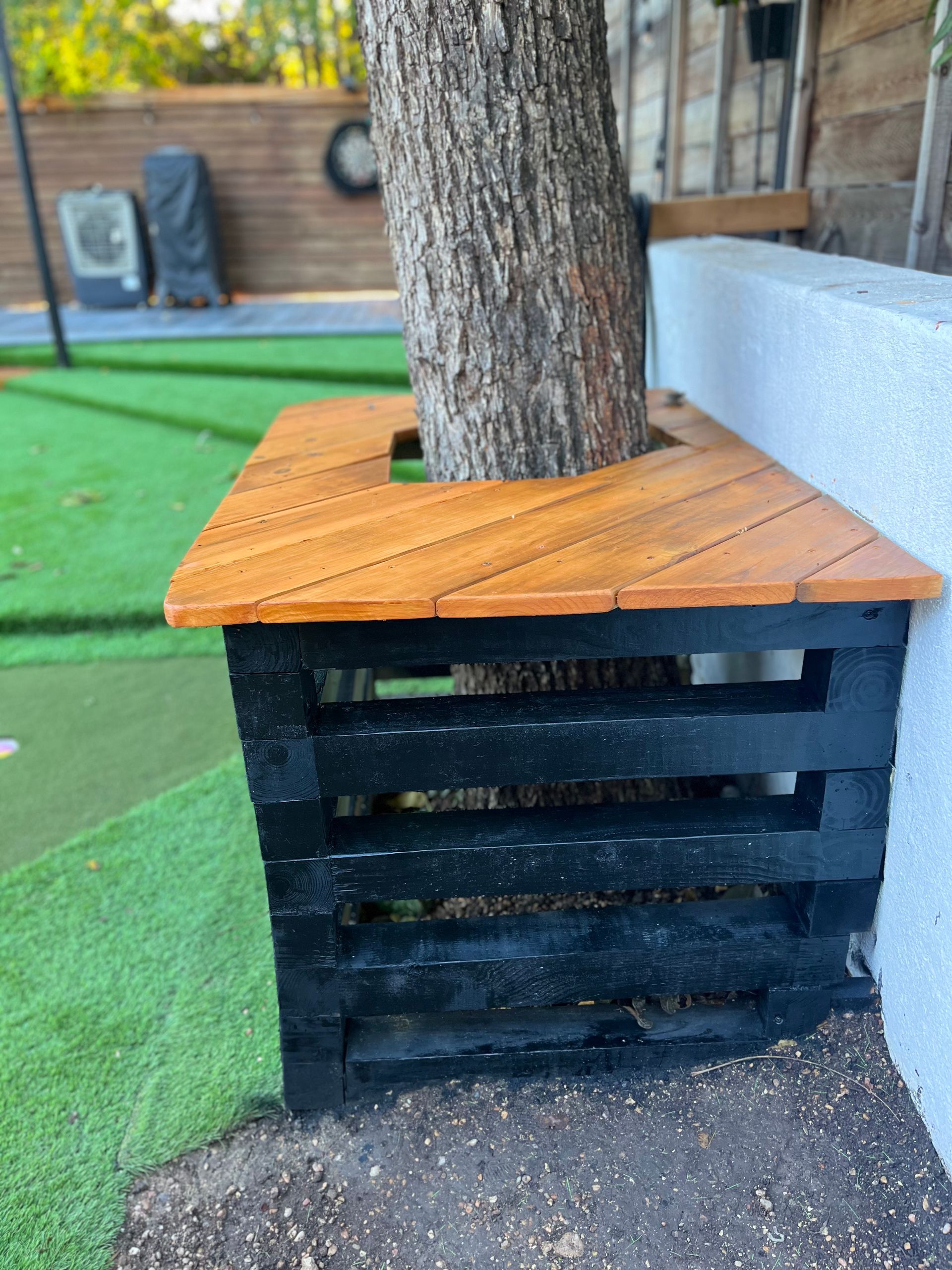 Tree trunk in a black wooden planter with an orange top on artificial turf beside a wall.