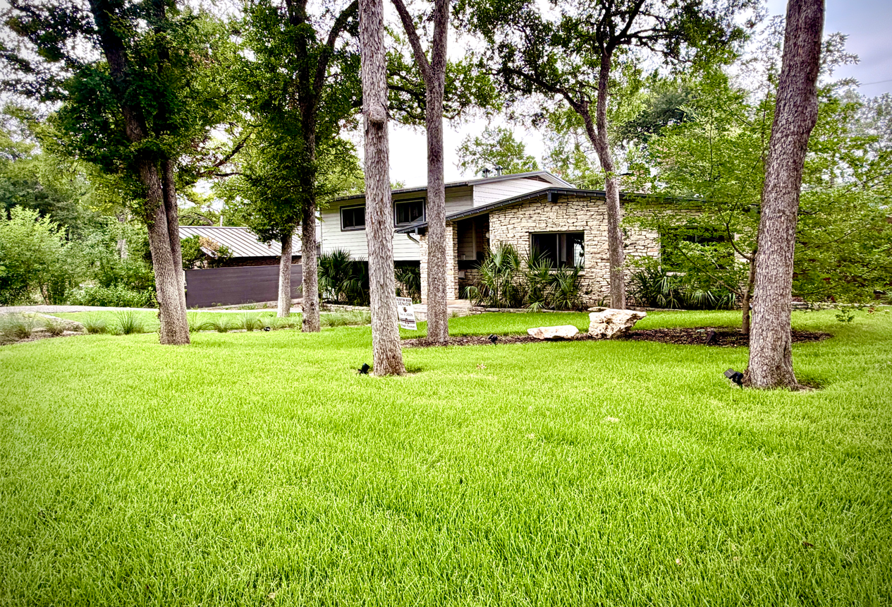 Backyard patio with green lawn, stone path, wooden deck, and tan fence beside a house.