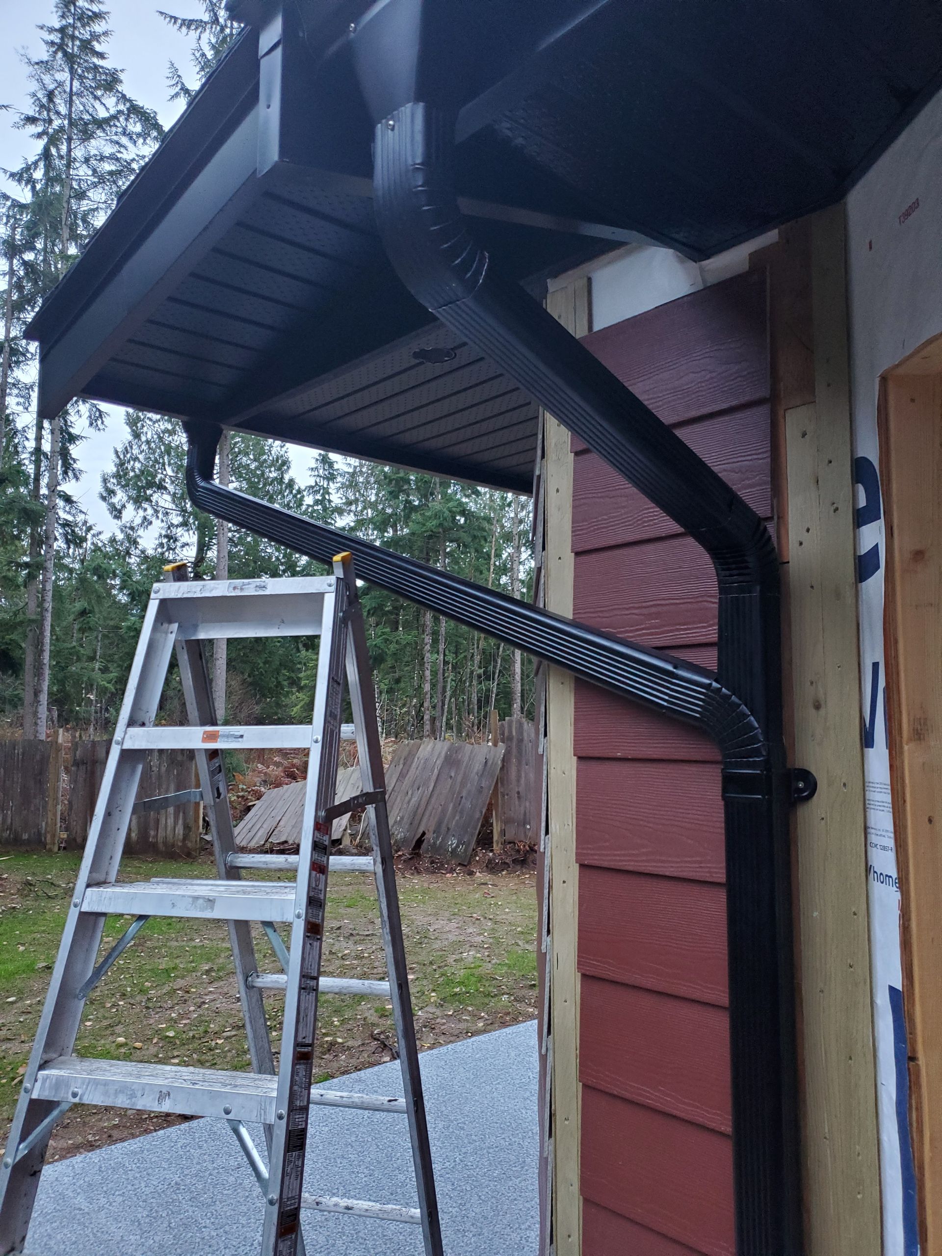 A black downspout extension attached to a house with red siding, positioned near an A-frame ladder on a gravel surface.