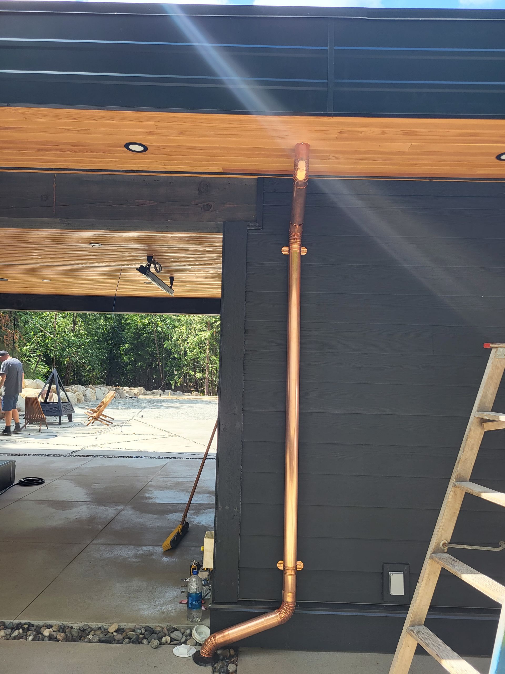 A copper downspout runs vertically along a dark-painted exterior wall, with an open patio area visible in the background.