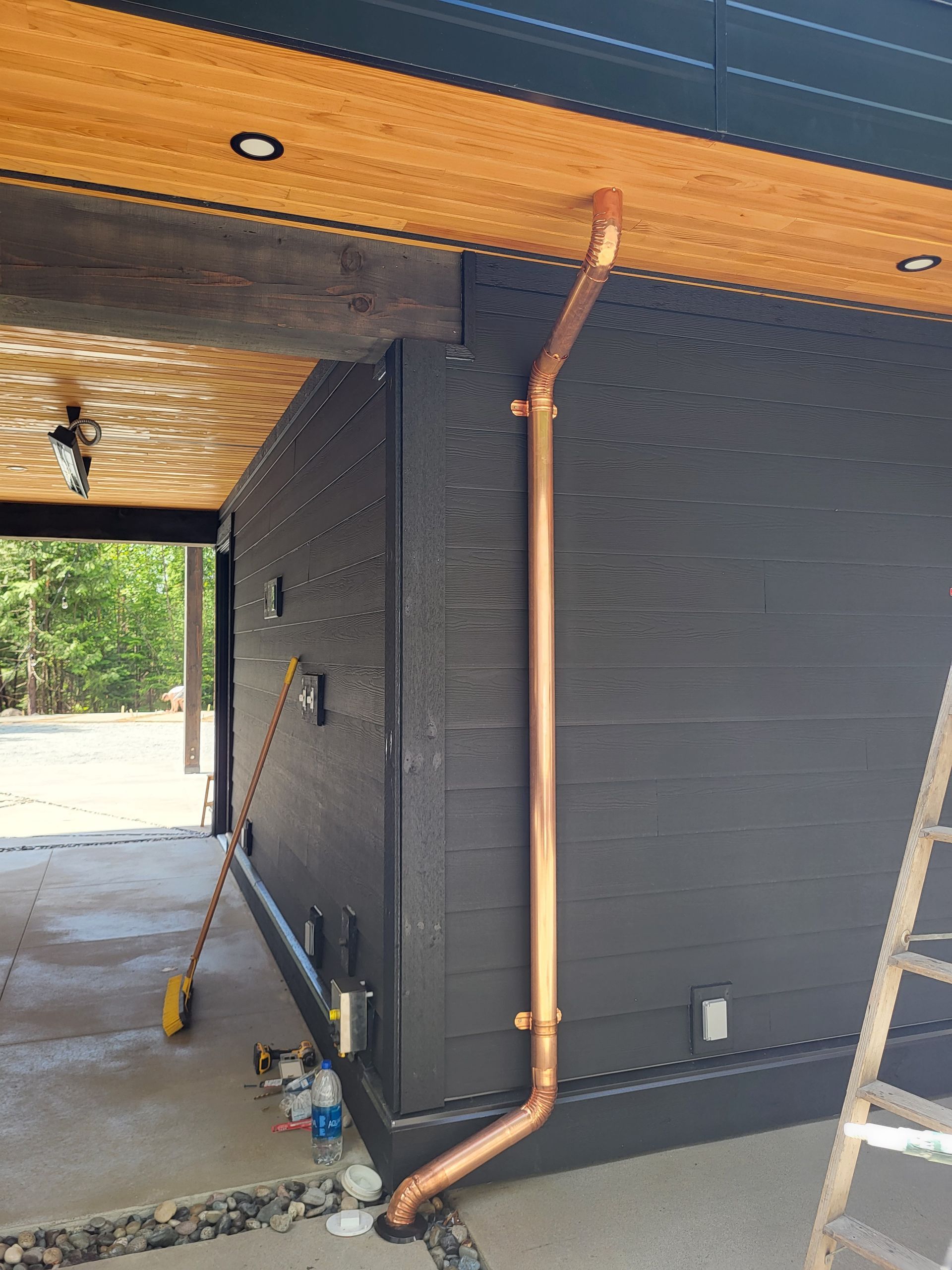 Copper downspout installed on the black siding of a modern house, with an open covered area and ladder visible.