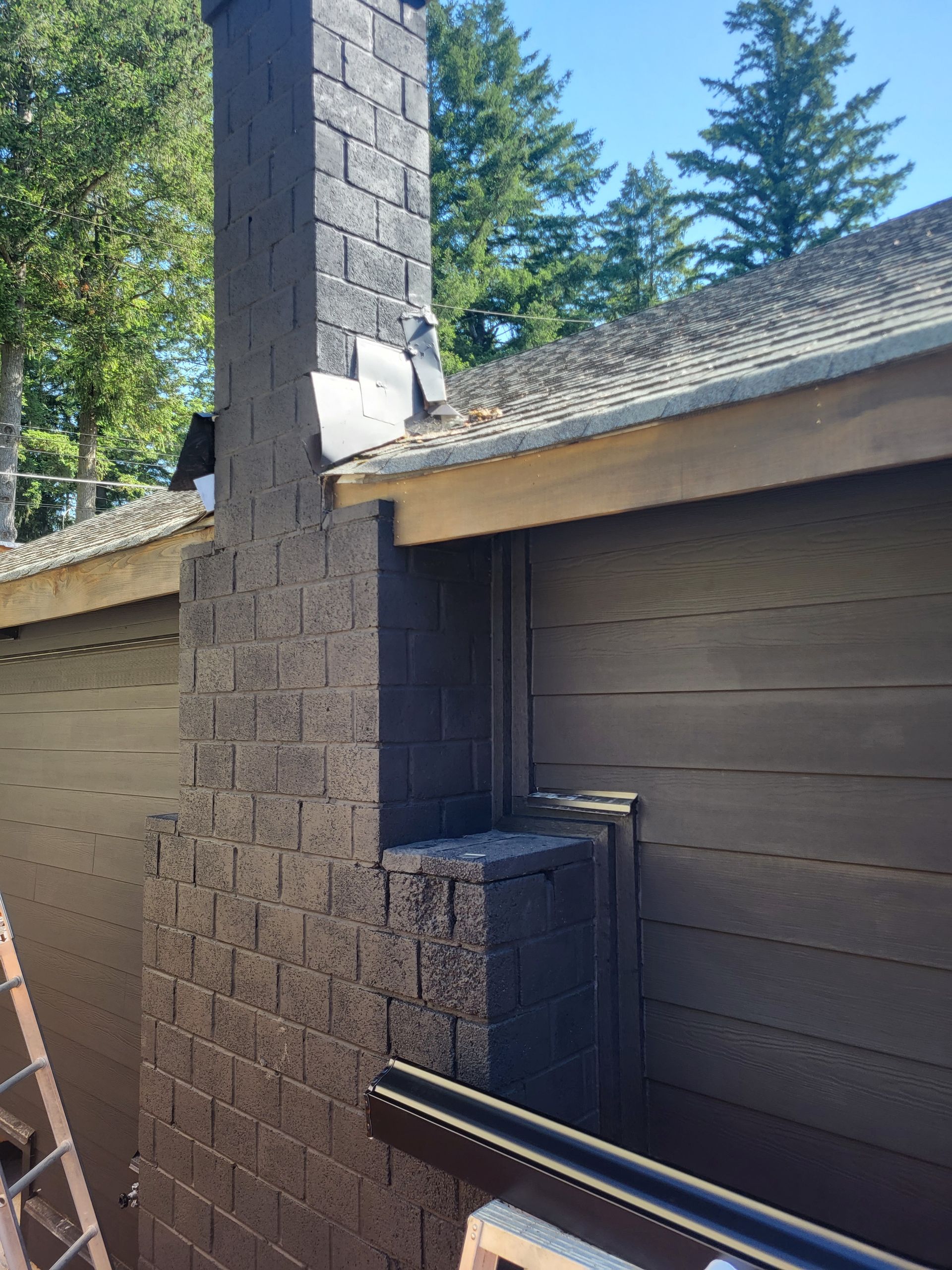 A dark-colored brick chimney rises from a roofline next to dark brown siding, showing visible flashing and exterior trim.