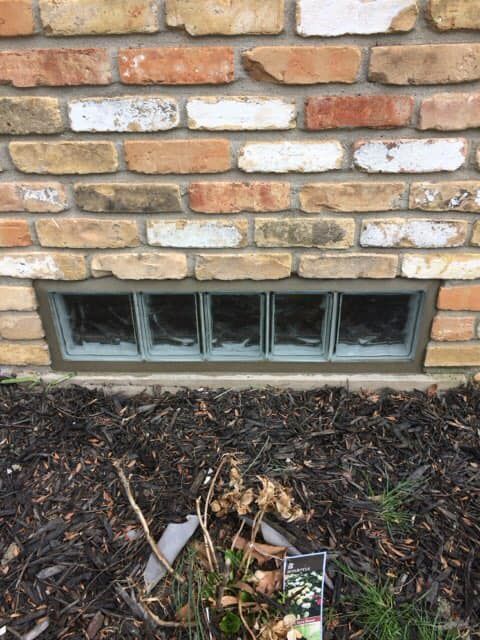 A rectangular glass block window set into a brick foundation wall above dark mulch.
