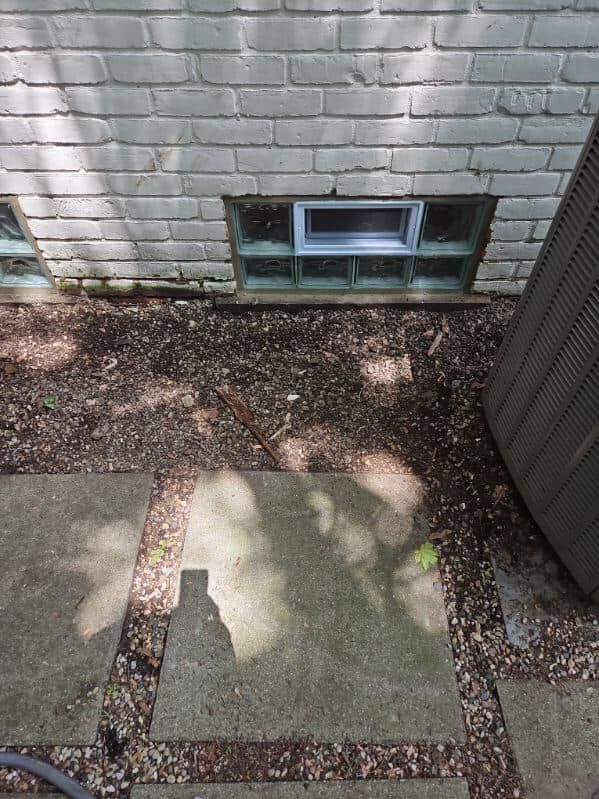 A white brick exterior wall with a glass block basement window, situated above a patio with scattered leaves.