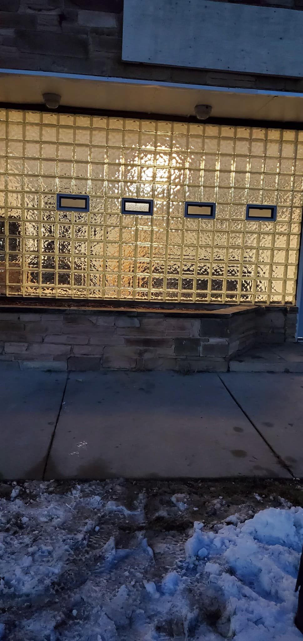 A sidewalk and stone wall in front of a building facade made of square glass bricks with four small, dark rectangular vents.