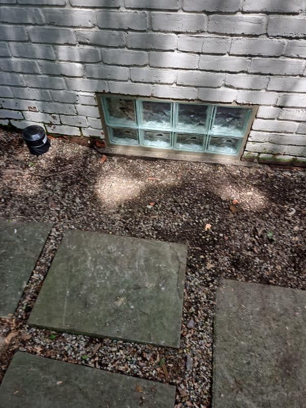 A small, glass-block basement window set in a white painted brick wall above a gravel path with stone pavers.
