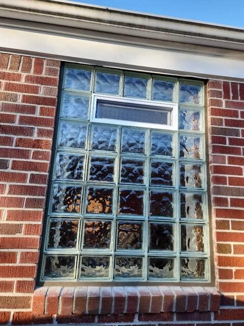 A rectangular window made of glass blocks, set in a red brick wall with a small horizontal sliding vent at the top.