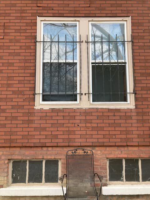 A brick building exterior featuring a dual-pane window with security bars above two smaller basement windows and a chair.