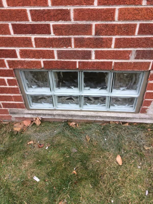 A glass block basement window embedded in a red brick wall above a patch of grass.