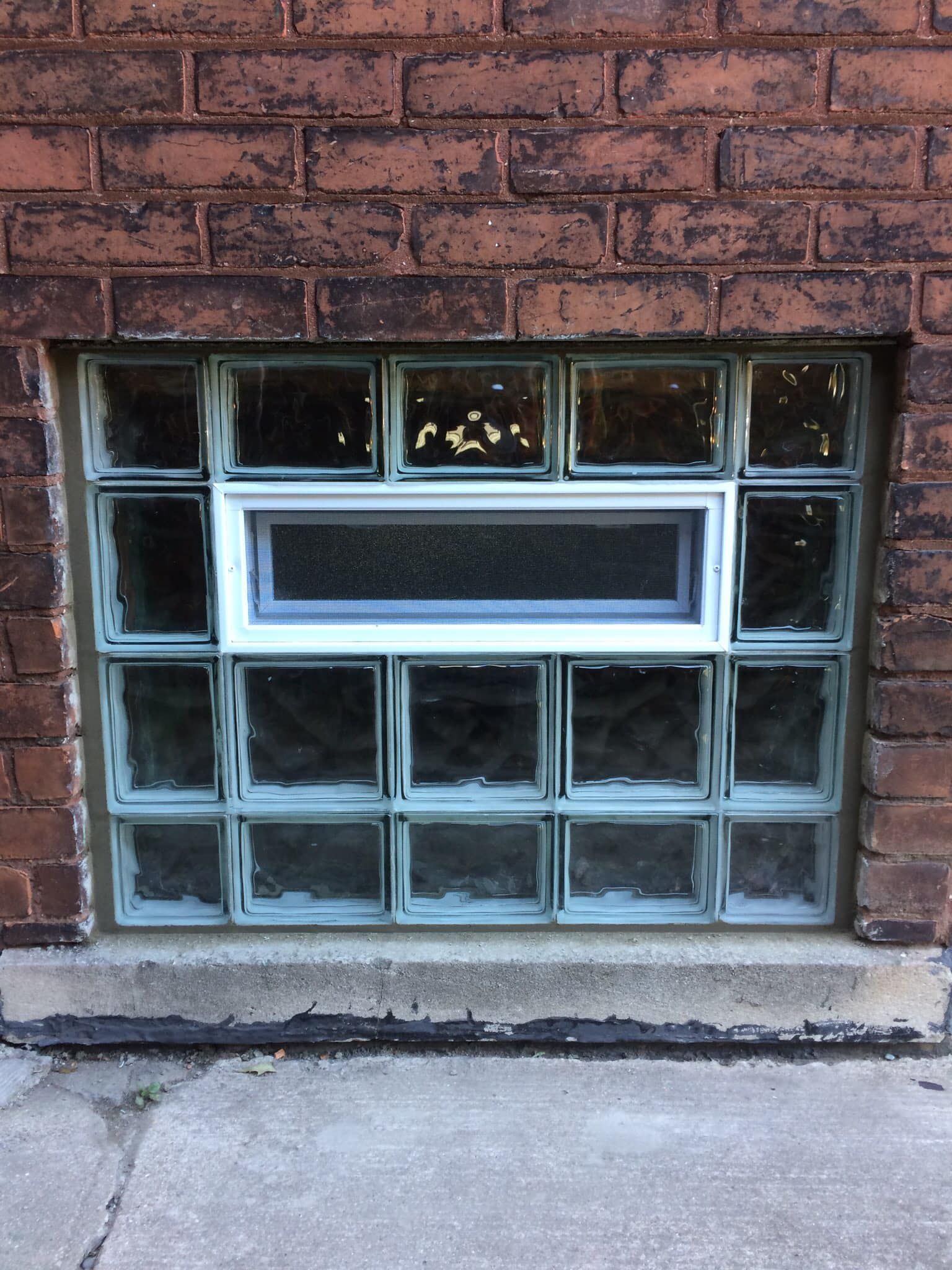 A rectangular basement window made of glass blocks with a central white-framed horizontal vent, set in a brick wall.