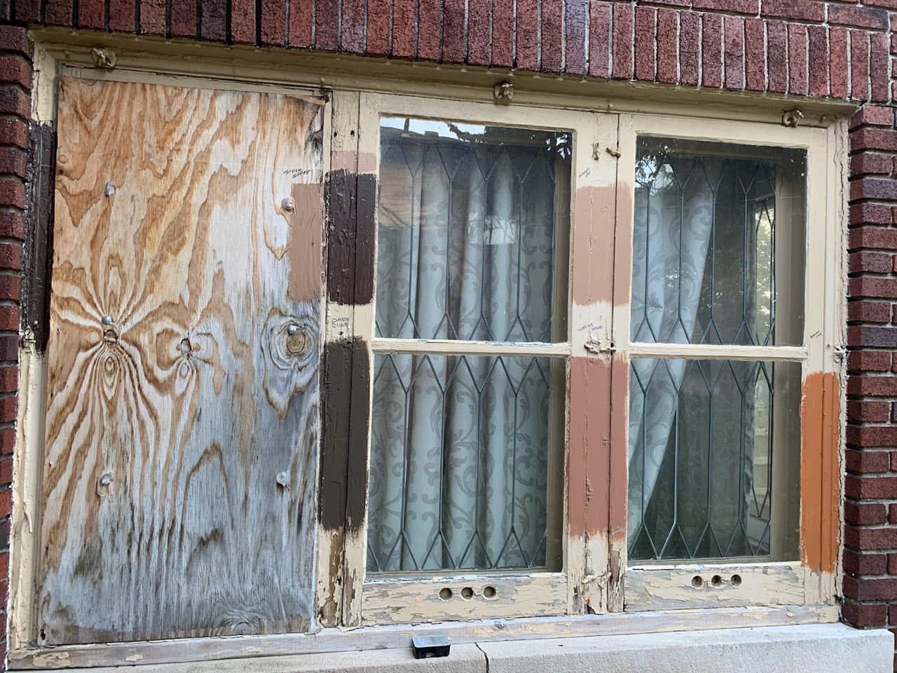 A window with one boarded-up pane and two glass panes, with several paint color test swatches applied to the wood frame.