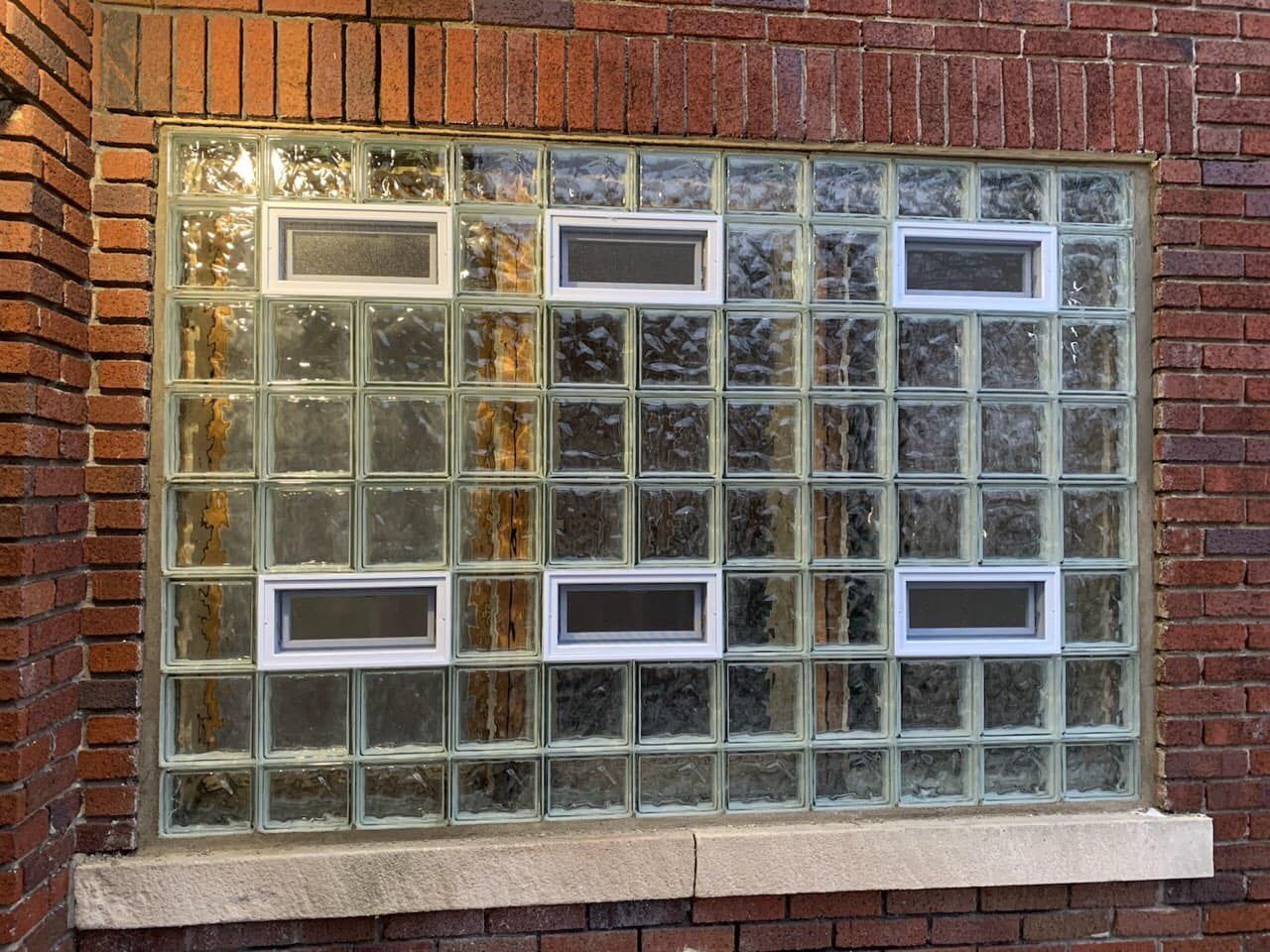 A brick wall containing a square glass block window with six small, rectangular, operable ventilation vents.