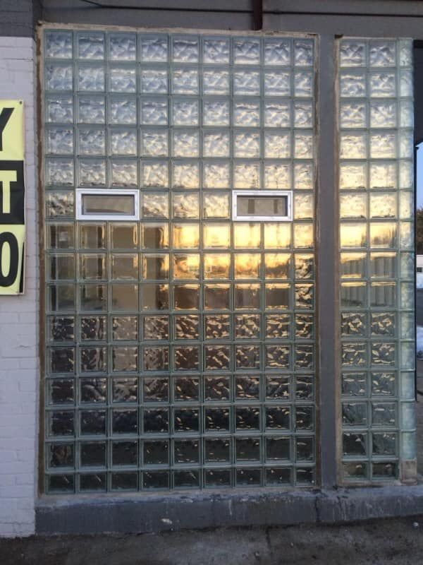 A large wall made of translucent square glass blocks, featuring two small, rectangular horizontal vents in the center.