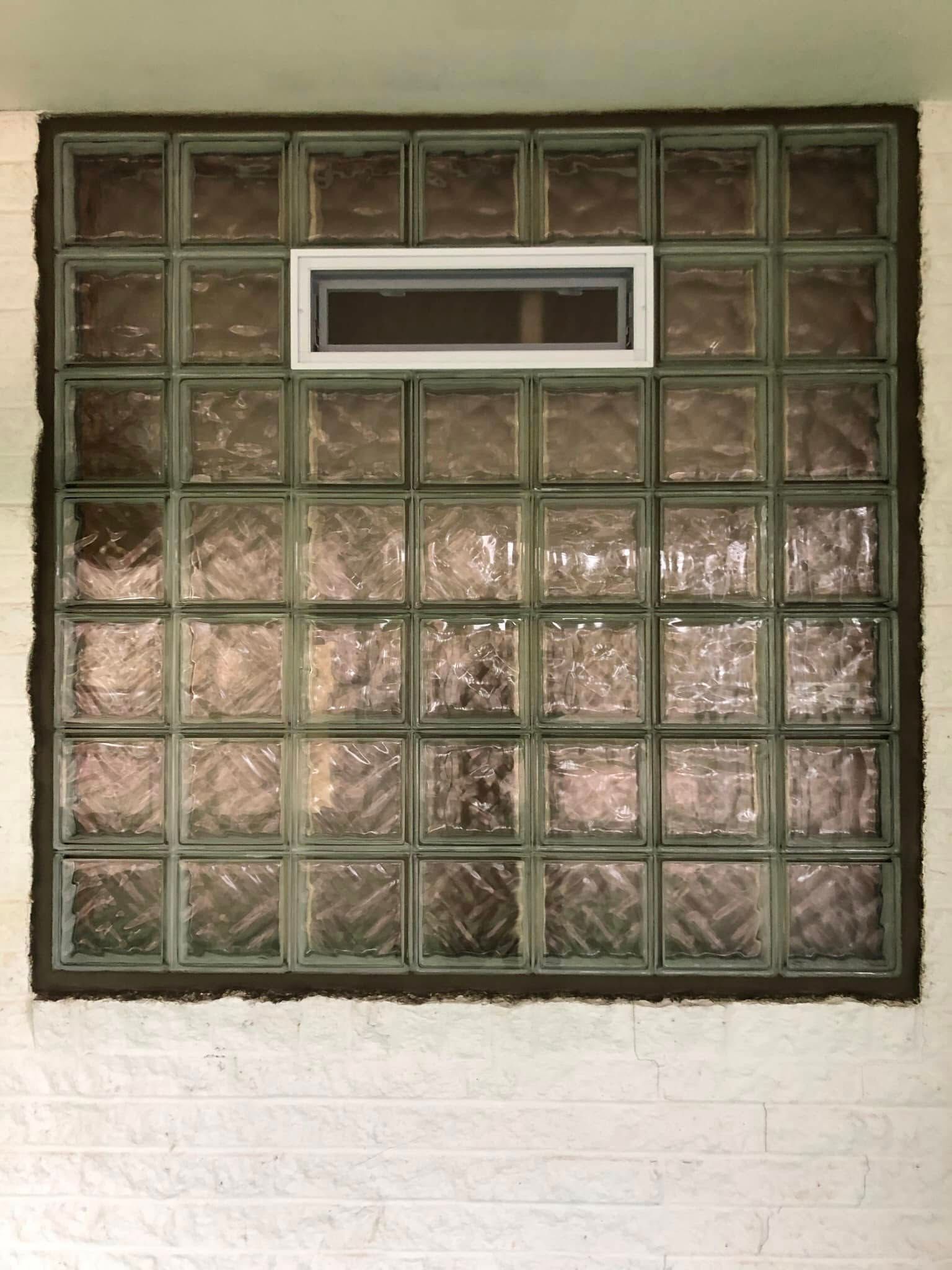A square glass block window set in a white brick wall, featuring a small, horizontal rectangular vent in the upper center.