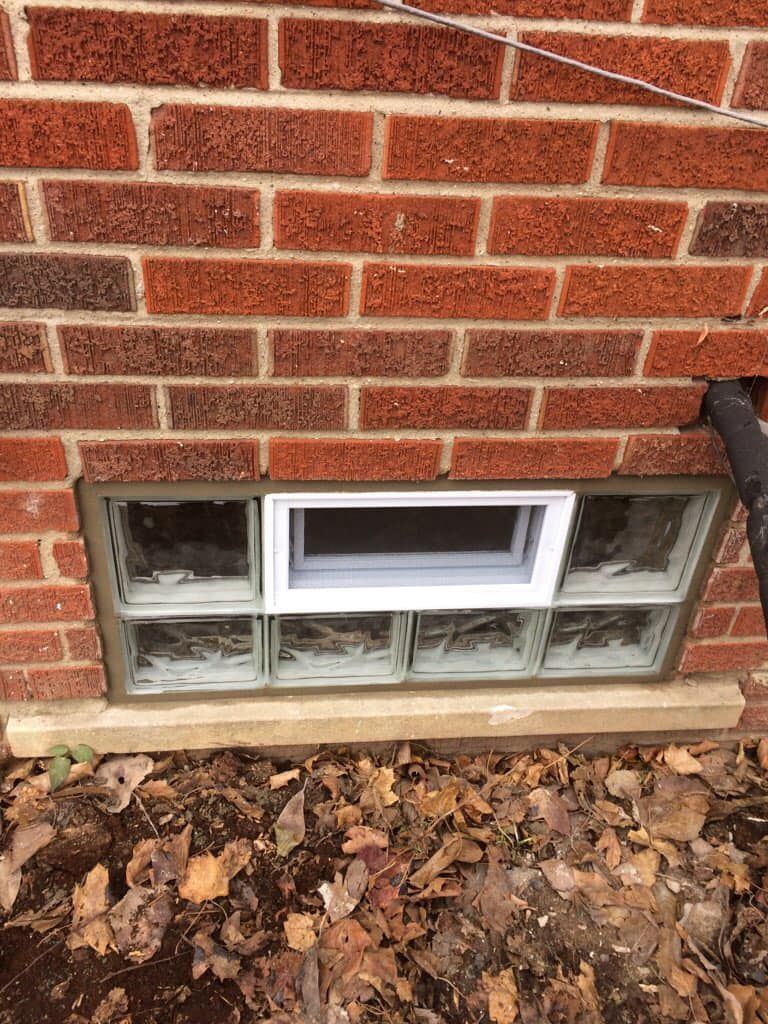 A glass block basement window in a red brick exterior wall with a central, small white-framed slider window.