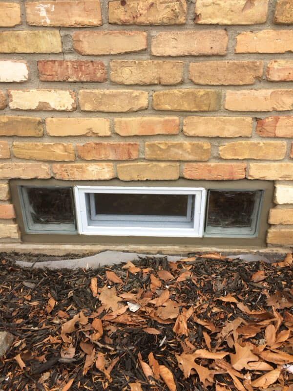 A rectangular basement window set in a light-brown brick wall above a garden bed with fallen leaves.