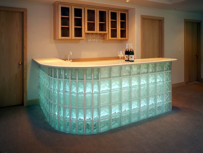 A curved home bar with a glowing, lit glass block front, white countertop, and light wood cabinetry in a finished room.