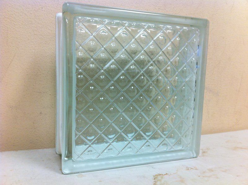 A clear glass block with a square grid pattern, resting on a flat surface against a plain wall.