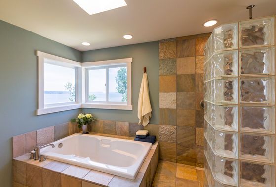 A bathroom with a white soaking tub, blue walls, a glass block shower, and corner windows with a water view.