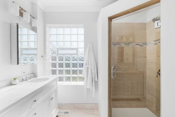 A bright, modern bathroom featuring a white vanity with a glass sink, a glass block window, and a tan tiled walk-in shower.