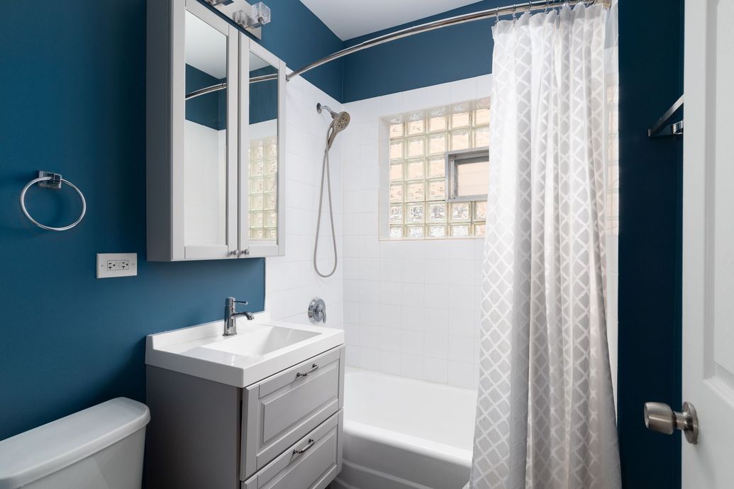 Modern bathroom with dark teal walls, white tiled tub-shower, glass block window, gray vanity, and white shower curtain.