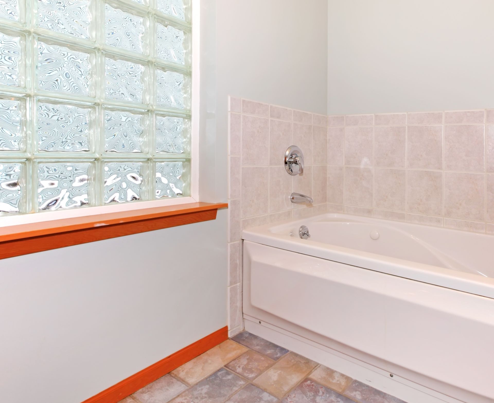 A white bathtub with tiled walls next to a glass block window with an orange-stained wood sill and floor trim.