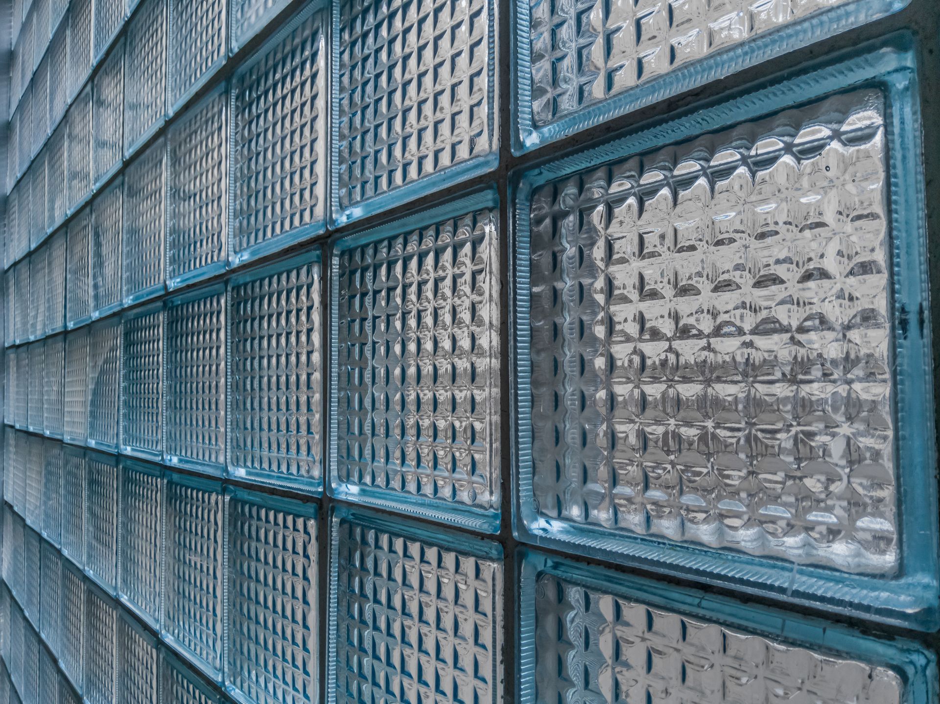 A close-up, angled view of a textured glass block wall with a repetitive grid pattern, illuminated by natural light.
