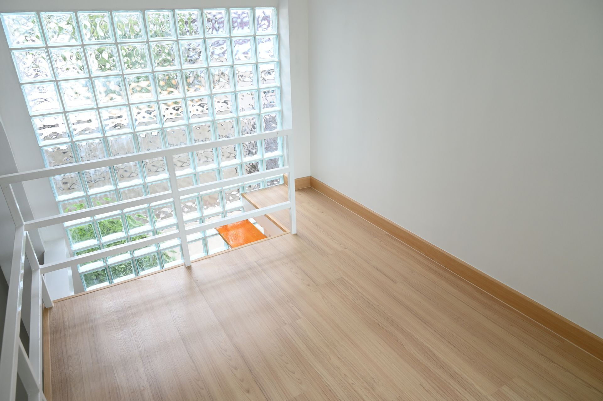 A bright room featuring light wood flooring, a white wall, and a large grid of glass blocks against a white railing.