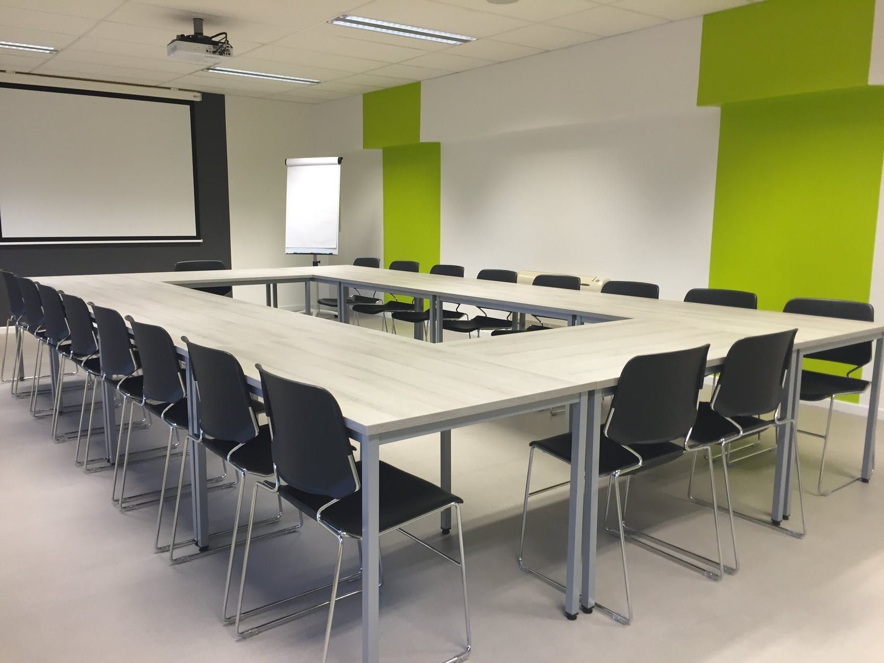 Empty conference room with U-shaped tables, black chairs, projector screen, and bright green accent wall
