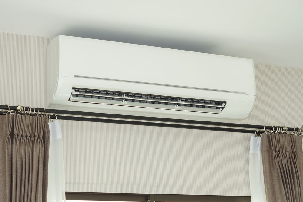 White Wall-Mounted Air Conditioner Above a Window With Brown Curtains — Inland Electrical in Humpty Doo, NT
