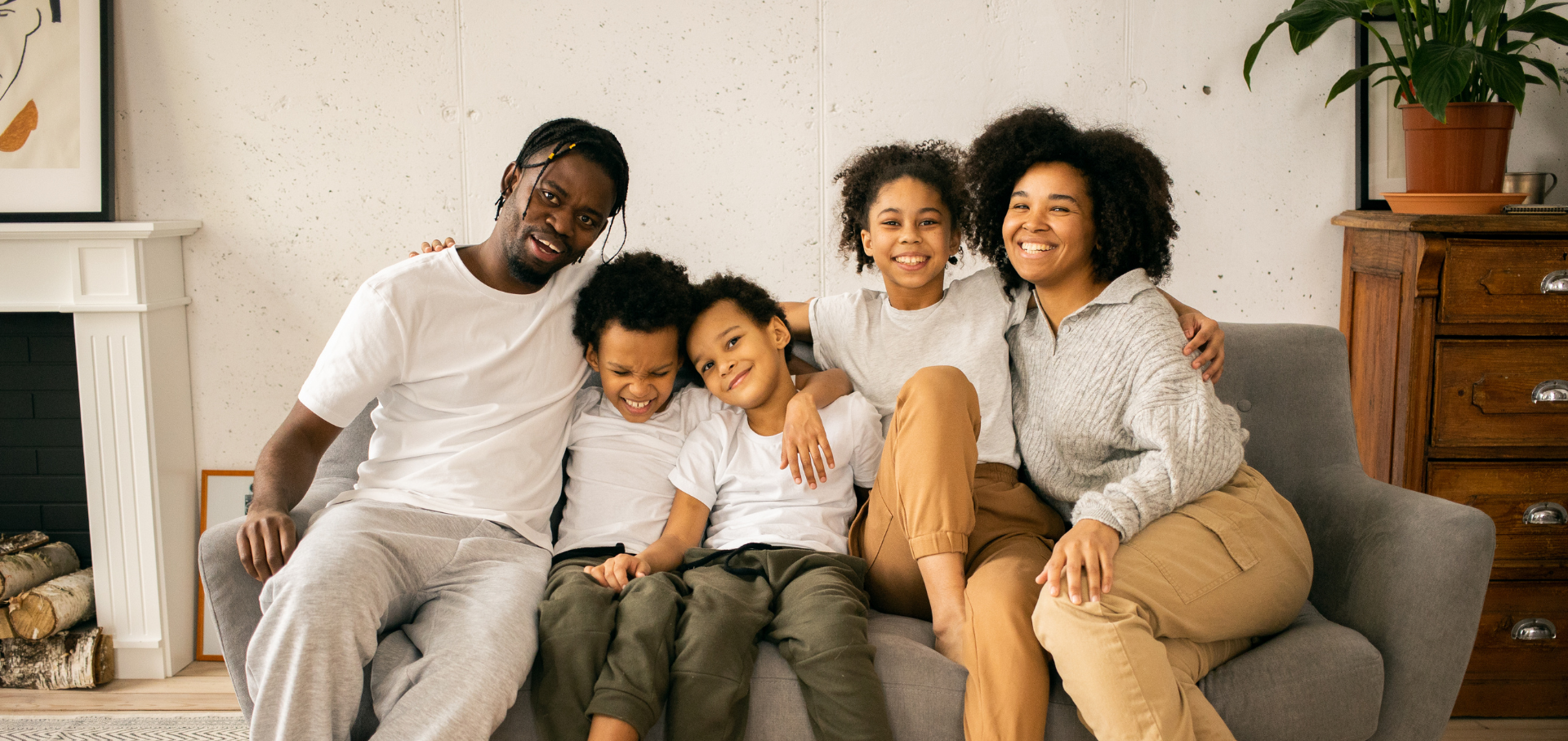 Family of five sitting on a gray couch, smiling. A father and mother hug their three children.