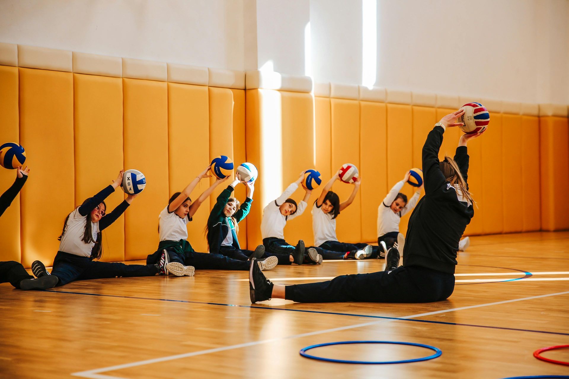 Children stretching with balls in a gym, led by an instructor, sitting on wooden floor.