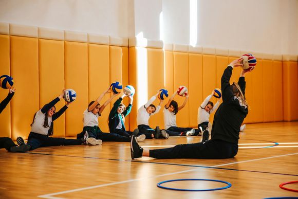 Students and instructor stretching with balls in a gymnasium, extending arms overhead while seated.