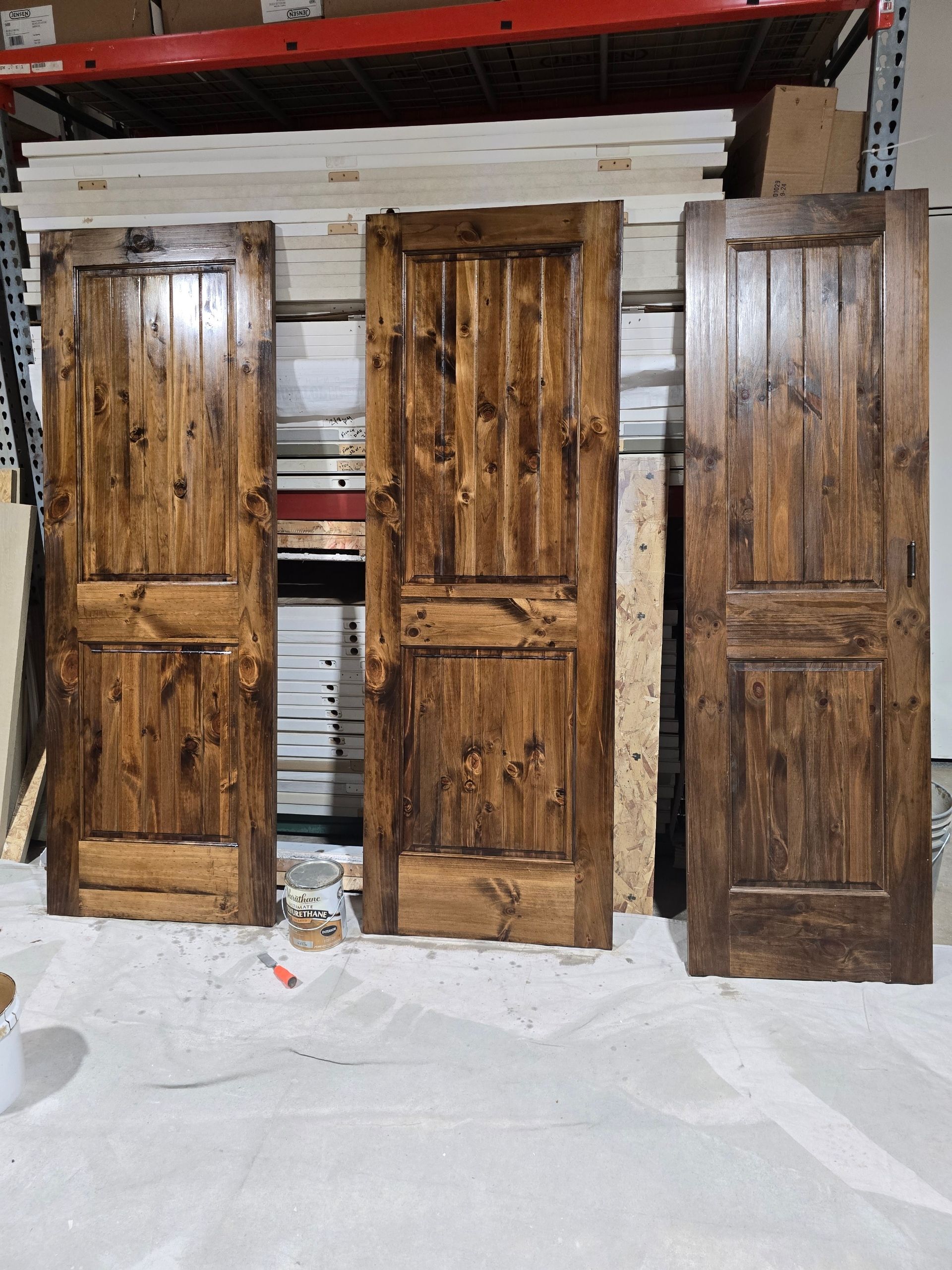 Three rustic wooden doors leaning upright in a workshop, with a red shelf in the background.