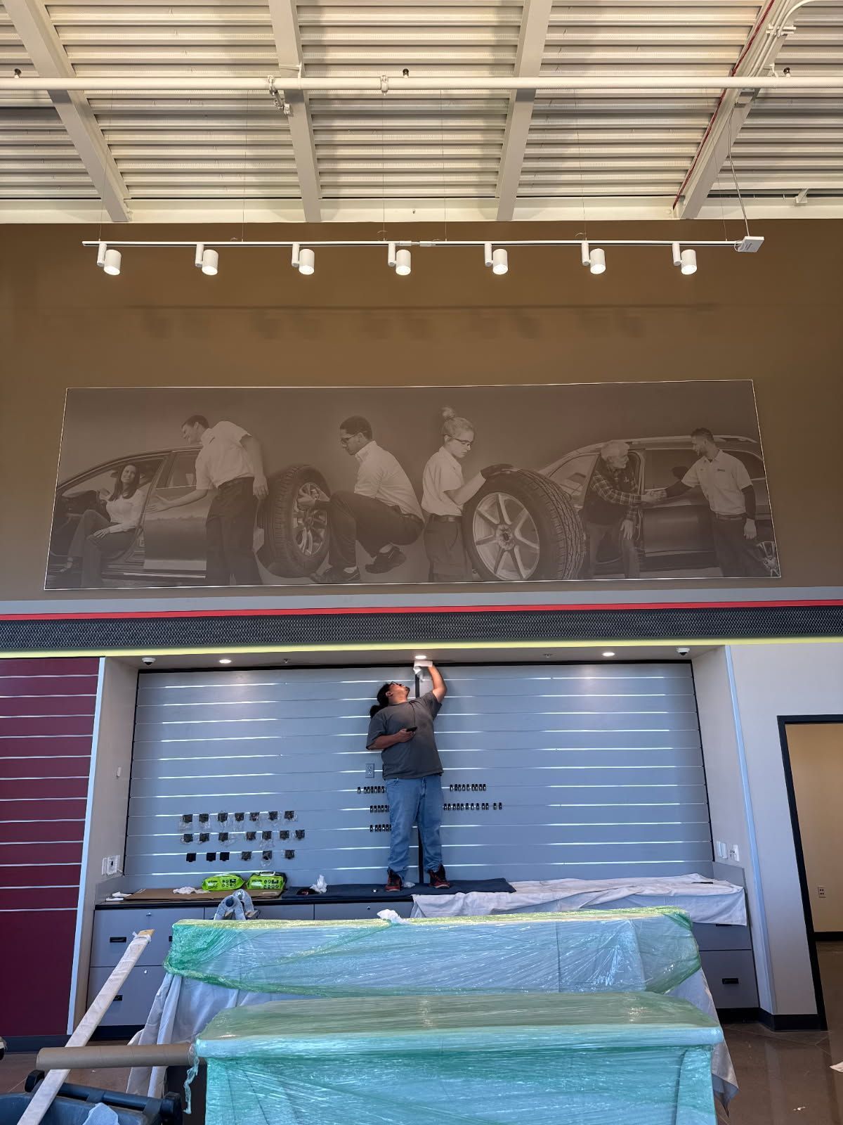 A person on a ladder installing something on a wall below a large mural of people with a car in a showroom.