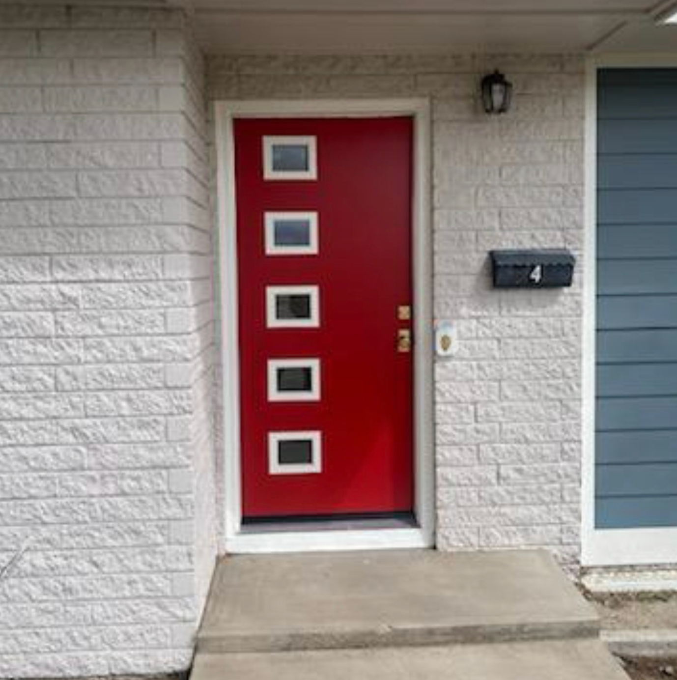 Red front door with four small windows beside a black house number plaque on a white brick entryway