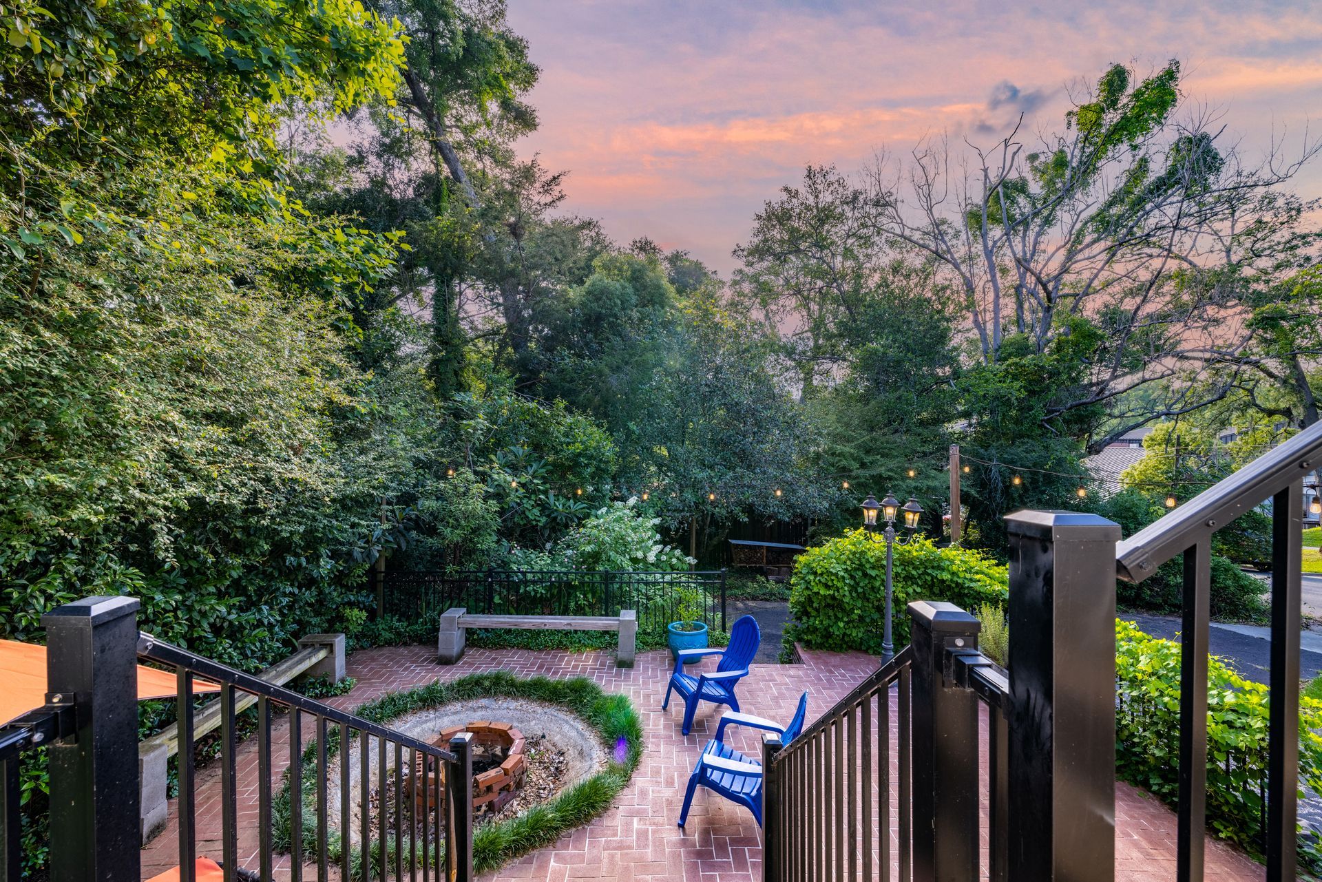 Outdoor patio with fire pit, blue chairs, brick and wrought iron. Green trees and sunset sky.