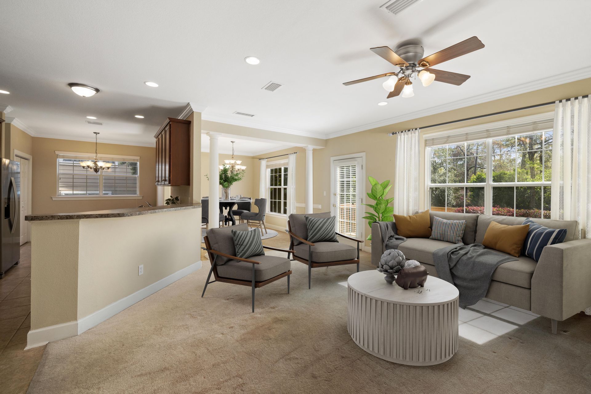 Spacious living room with tan carpet, neutral walls, open to kitchen and dining area; light-filled with large windows.