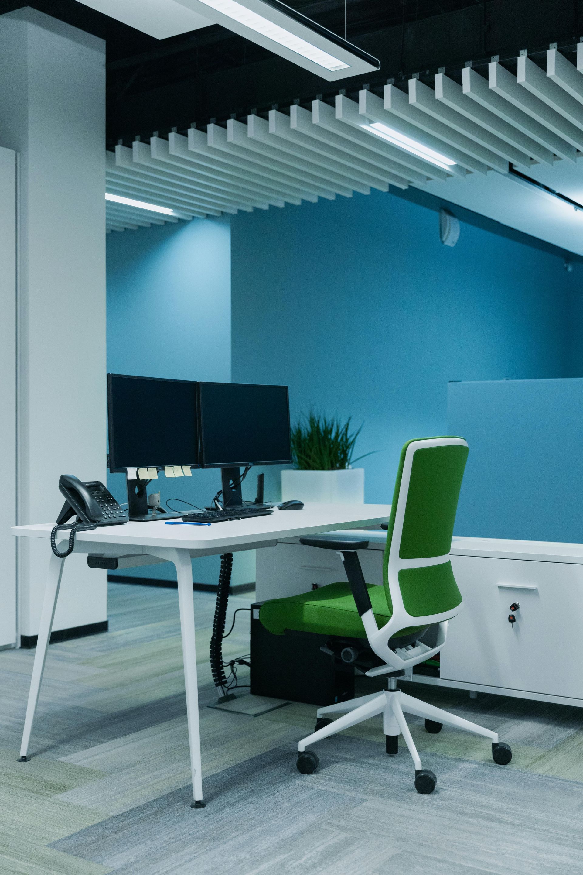 Modern office workspace with white desk, dual monitors, and a green swivel chair against blue walls